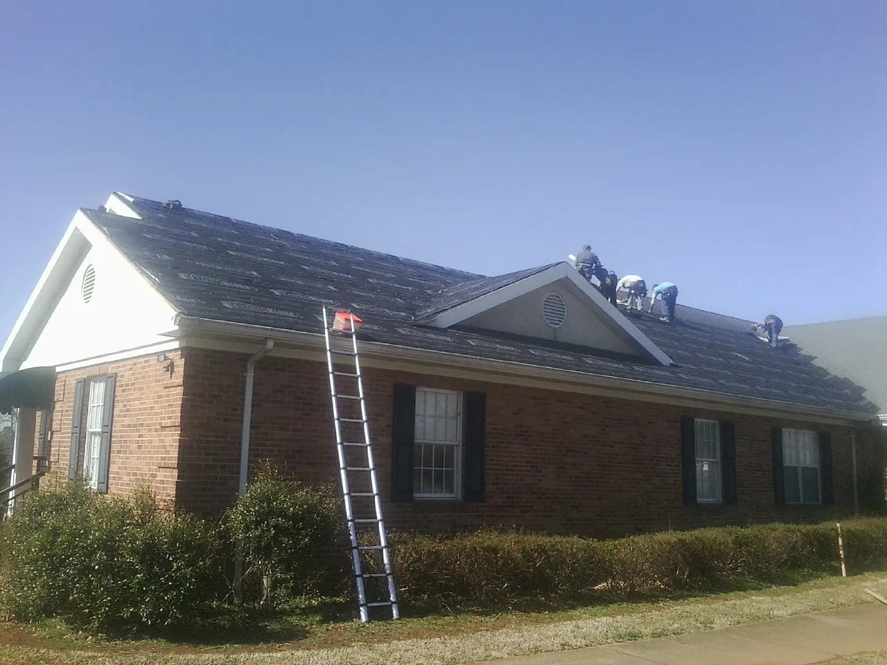 Crew tearing shingles off a brick ranch roof with a ladder set — Emergency Roof Repair work by our crew in Bombay