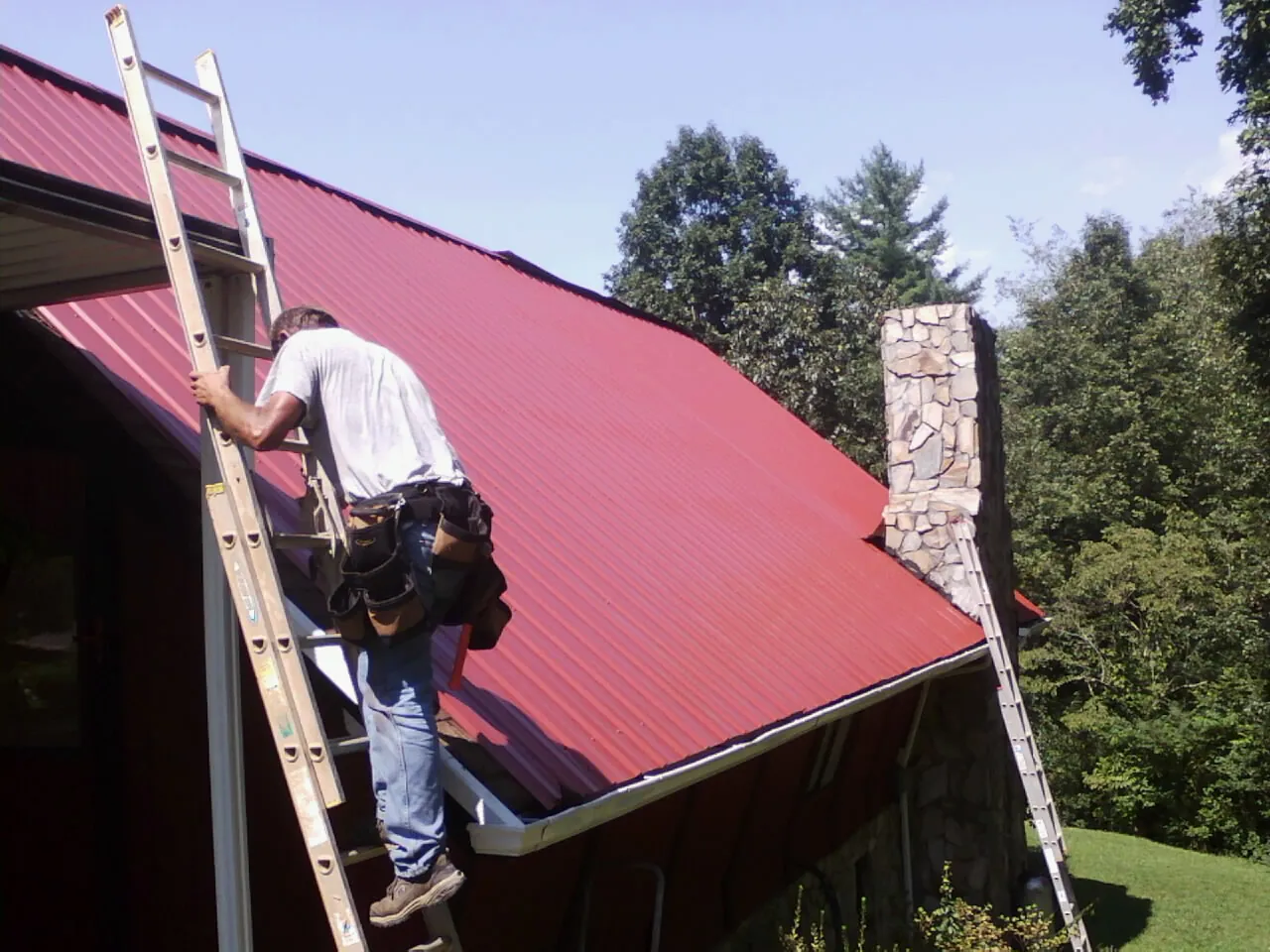 Roofer on a ladder beside a red metal roof and stone chimney — Soffit & Fascia Repair by Roofing Brothers in Bombay